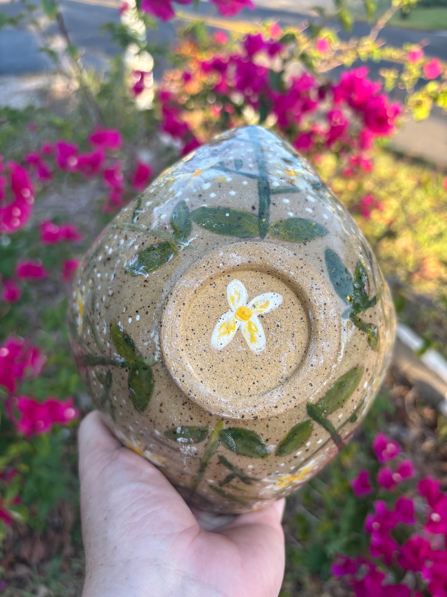 Photo of the bottom of a speckled pottery bowl painted with white and yellow rue flowers. The bottom of the bowl has a hand painted blossom in the foot ring. The bowl is held in the artist's hand and set against a background of bushes with pink flowers.