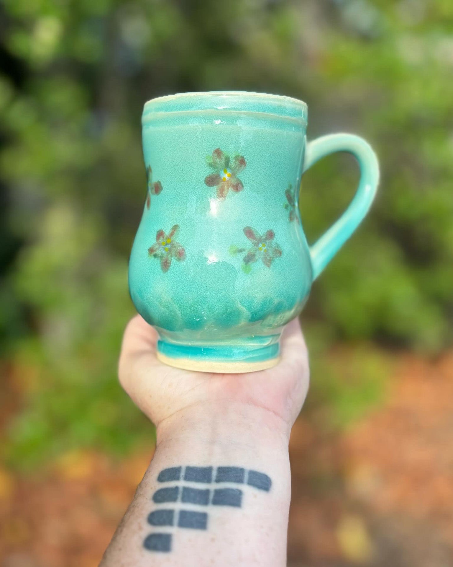 Photo of a large curvy pottery mug in a blue green glaze with red painted flowers.  The mug is held in the artist's hand and is set against a backdrop of green leaves.