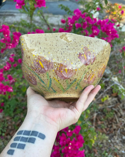 Photo of a large speckled ceramic bowl painted with pink and yellow tulips.  The bowl is held in the artist's hand and set against a bush with pink flowers.