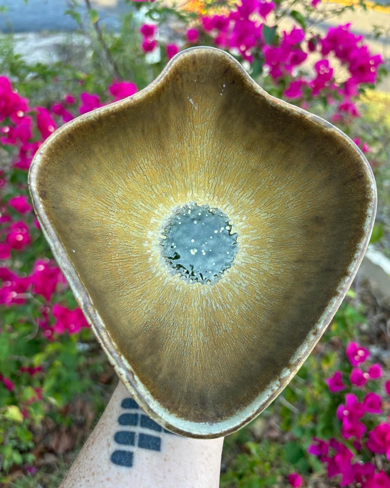 Photo of a yoni-shaped ceramic bowl with buttery glaze that runs elegantly down to the center of the bowl.  In the middle, a glassy green pool shines in the sun.  The bowl is held in the artist's hand and set against a background of bushes with pink flowers.