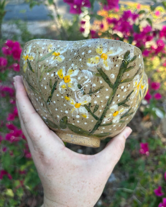 Photo of a speckled pottery bowl from the side.  The bowl is painted with white and yellow rue flowers on green stems with leaves.  white and dark brown flecks pop all over the bowl.  The bowl is held in the artist's hand and set against a green bush with pink flowers.