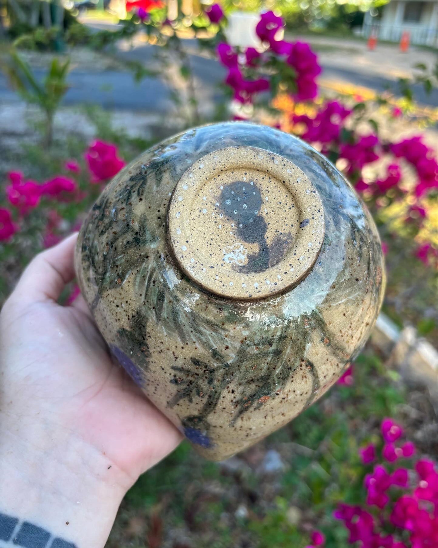 Photo of the bottom of a speckled ceramic bowl painted with purple thistle flowers on the outside and glazed with a deep purple on the inside.  The bowl is being held in the artist's hand against a blurred background of a green bush with pink flowers.