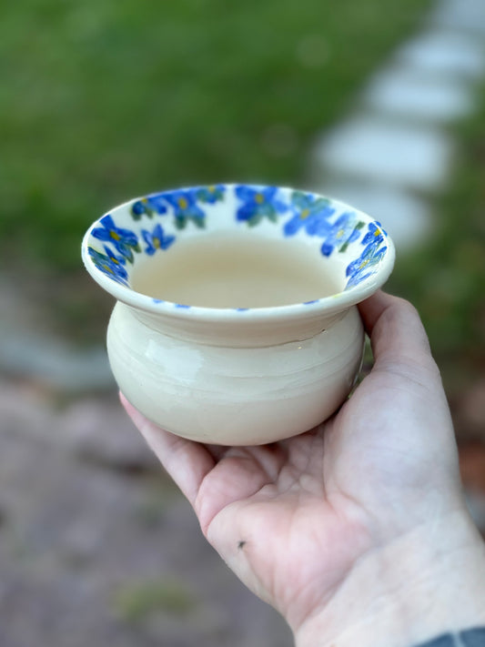 Photo of a white wheel thrown pottery vase held in the artist's hand.  The pot has a rounded bottom and wide, flared rim.  Blue flowers are hand painted around the rim of the pot. 