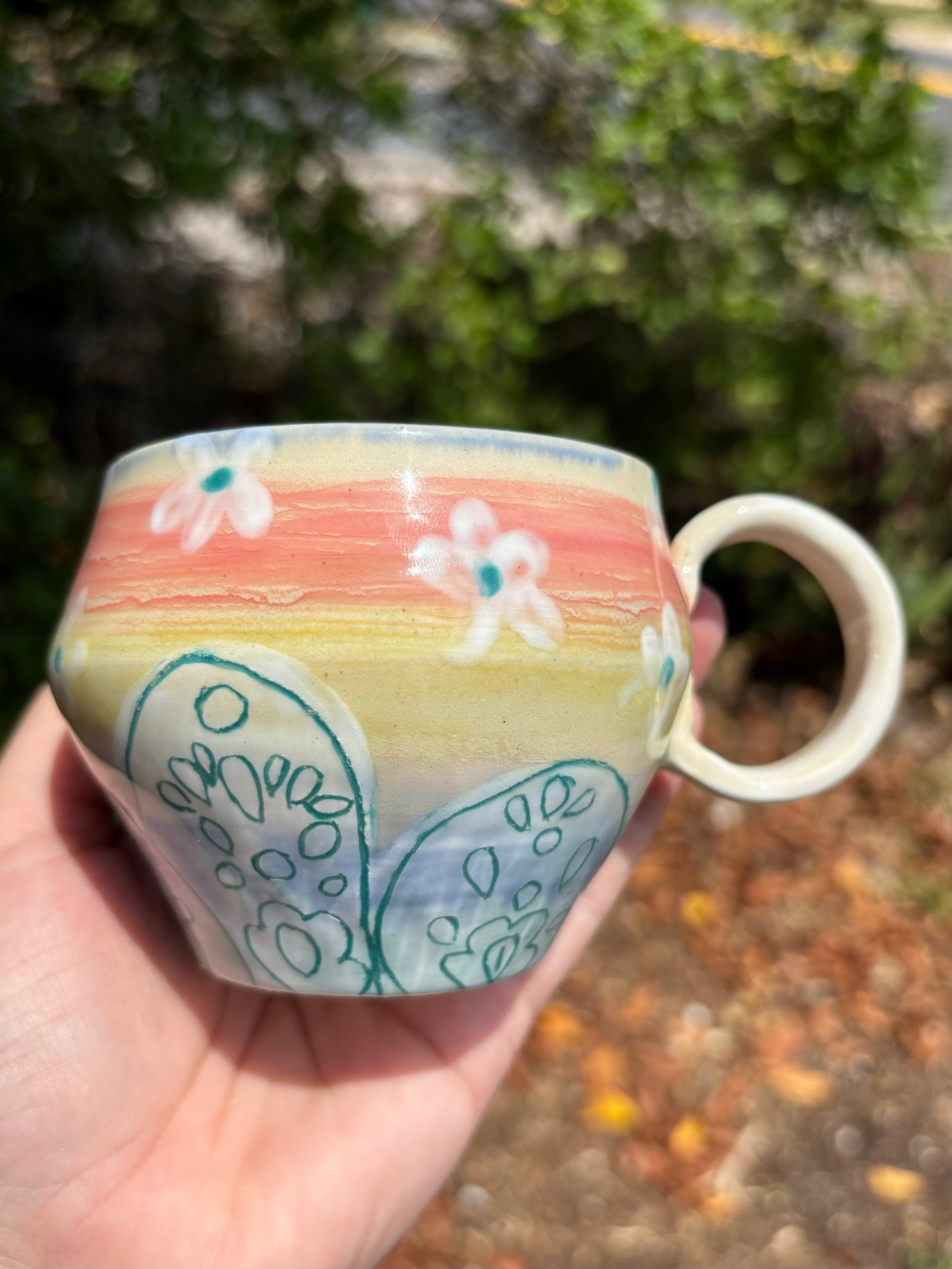 Photo of a wide mug with a round handle.  The mug is painted in a rainbow gradient and has a white petal design with teal inlaid zentangles on the petals.  There are white and teal blossoms painted around the body of the mug.