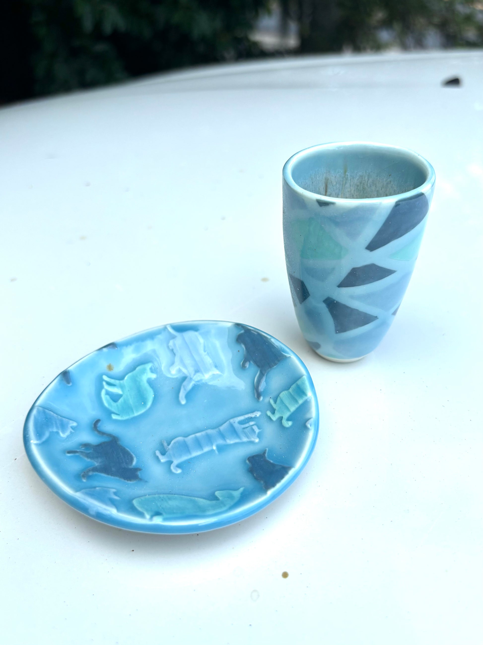 Photo of a small dish and matching cup in shades of blue. The dish has textured cats pressed into the clay, and the cup has stripes and lines that look like a wrapped up ball of yarn.