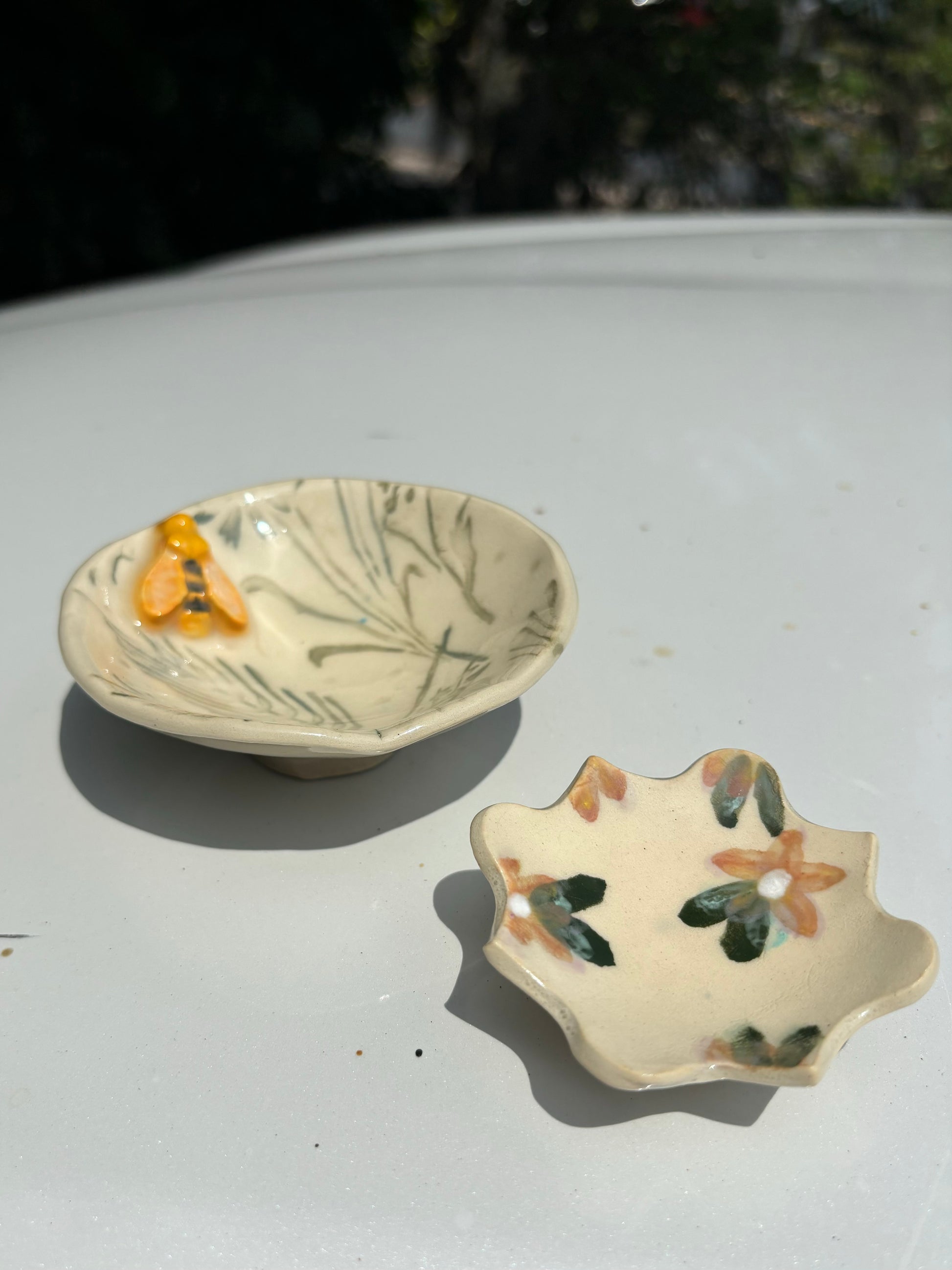 Photo of two small dishes featuring bees, green inlay grasses, and orange florals on a creamy buff clay body.