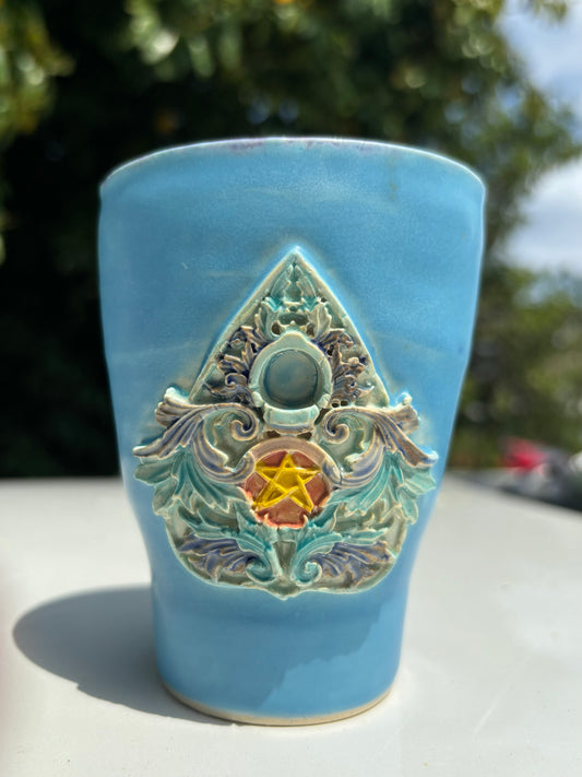 Photo of a tall blue ceramic mug with a hand painted ornate planchette decal attached to the front of the mug.  The planchette is ornate and filigree, painted in jewel tones with blues, purples, golds, and pinks.