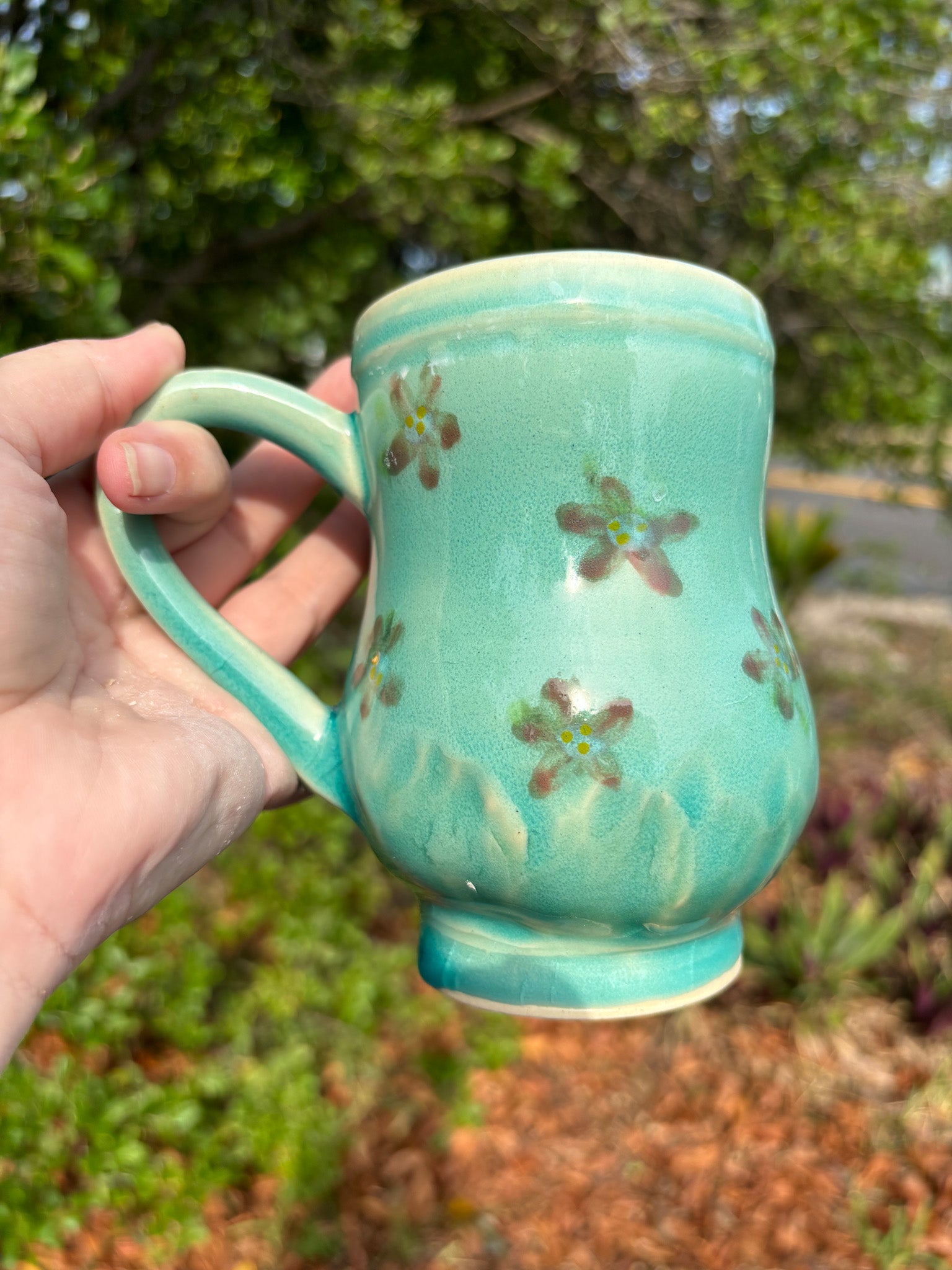 Photo of a large curvy pottery mug in a blue green glaze with red painted flowers. The mug is held in the artist's hand and is set against a backdrop of green leaves.