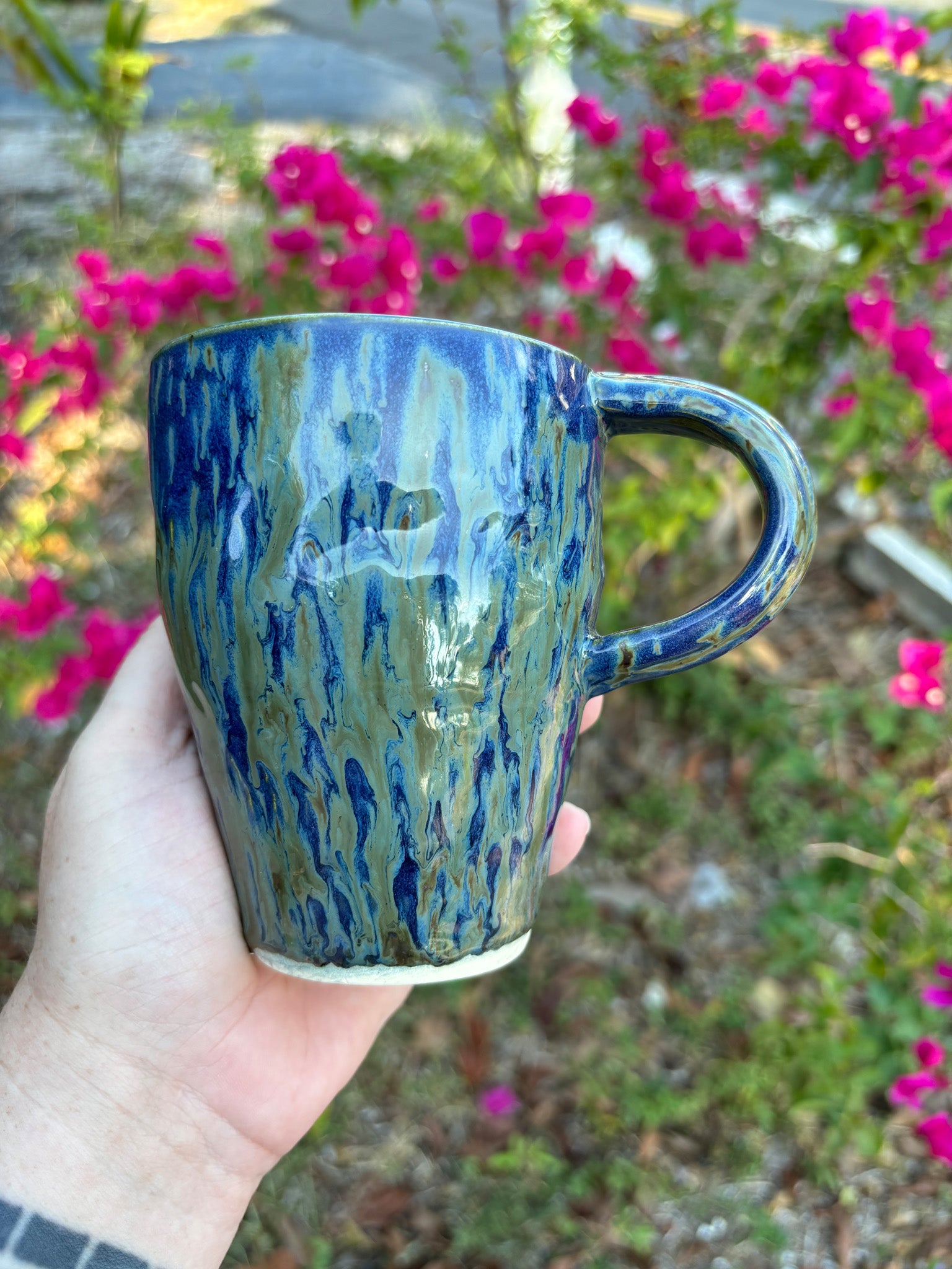 Photo of a tall pottery mug with a high modern handle.  It's glazed in a deep cobalt blue with streaks of greens like falling rain.  The mug is held in the artist's hand and sits against a background of green bushes with pink flowers.