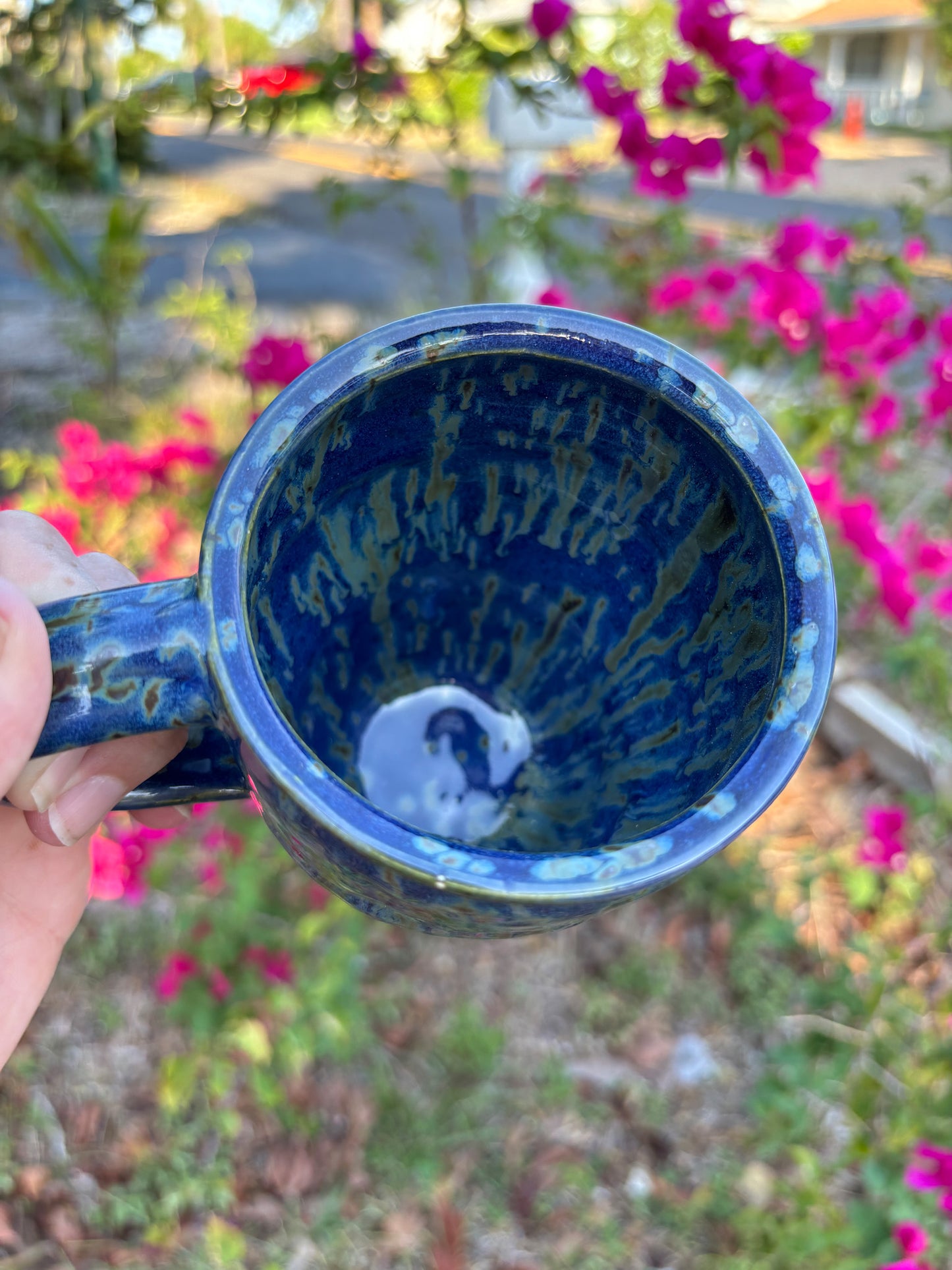 Top view of a cobalt blue pottery mug with green streaks like falling rain.  The streaks pool down the inside walls of the cup.  The cup is held in the artist's hand, set against a background of bushes with pink flowers.