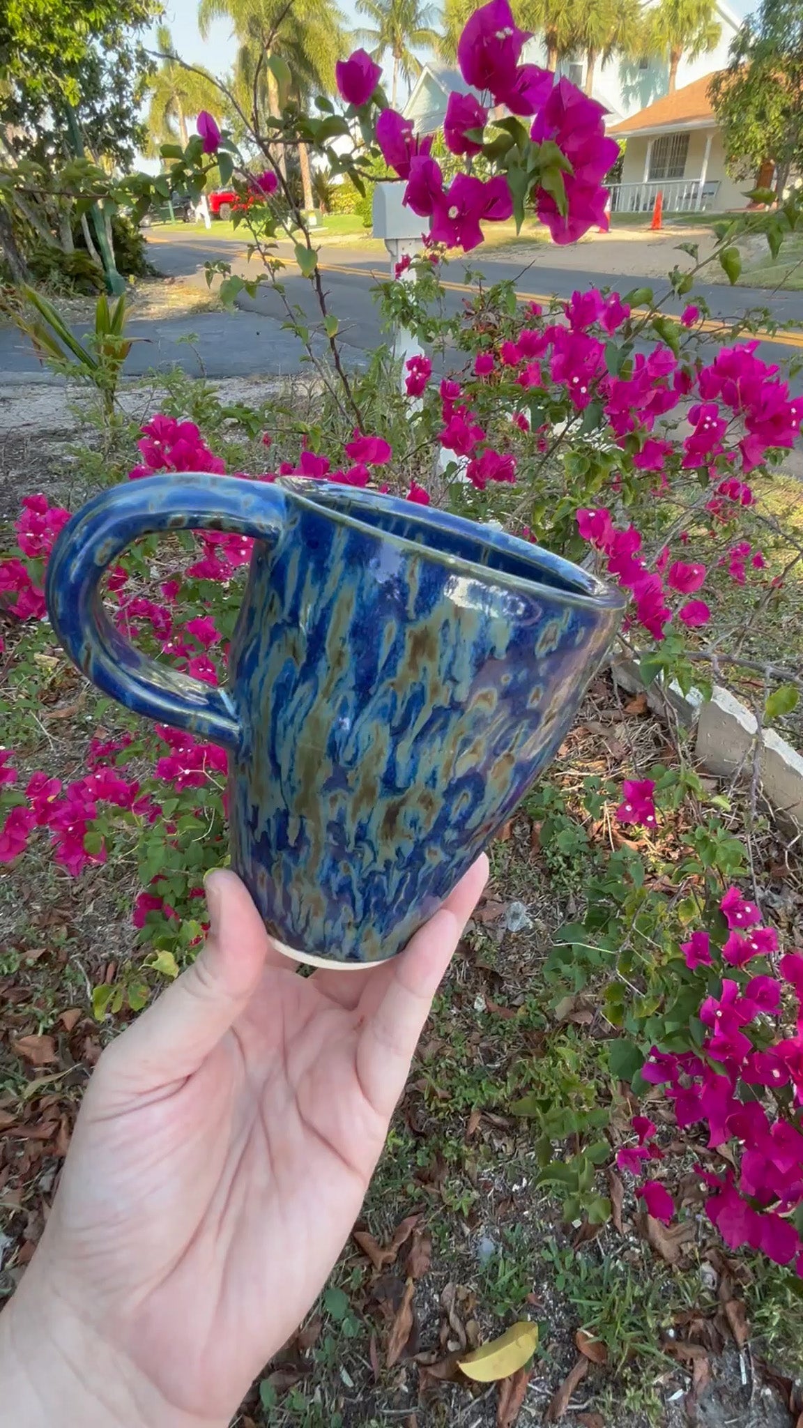 Photo of a tall pottery mug with a high modern handle. It's glazed in a deep cobalt blue with streaks of greens like falling rain. The mug is held in the artist's hand and sits against a background of green bushes with pink flowers.