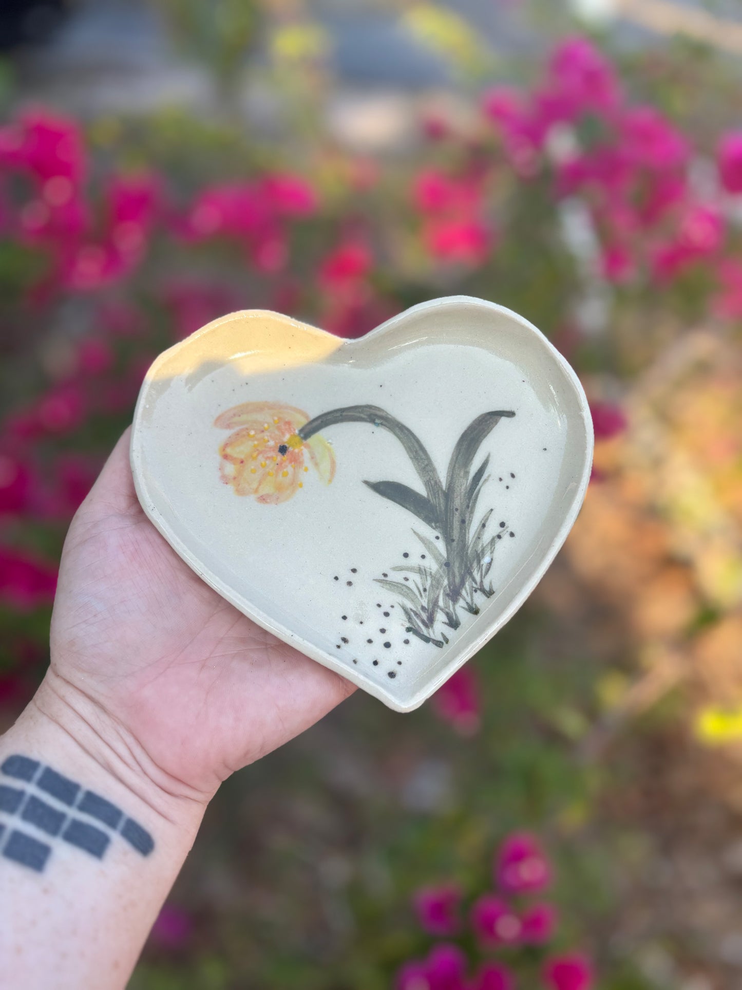 Photo of a heart shaped dish with a single yellow flower amid blades of grass.  The dish is held in the artist's hand and set against a background of bushes with pink flowers.