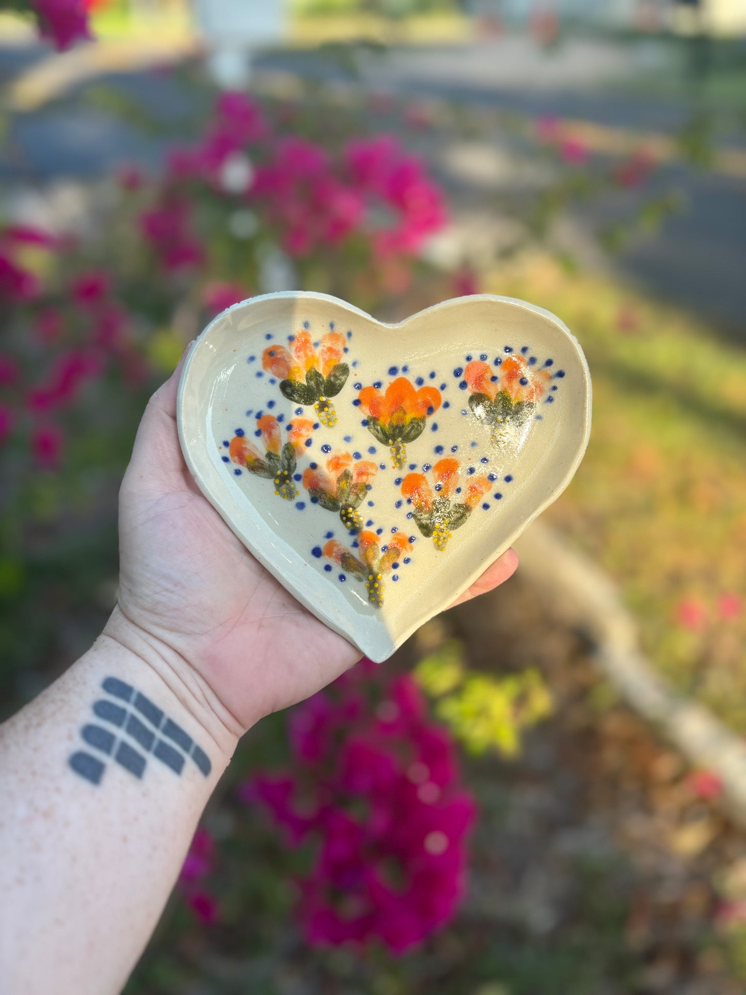 Photo of a heart shaped dish with vivid orange tropical florals and blue dots.  The dish is held in the artist's hand and set against a background of bushes with pink flowers.