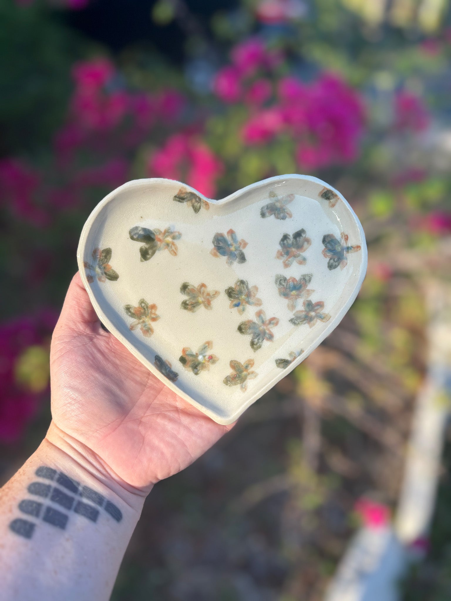 Photo of a heart shaped ceramic dish with hand painted flowers in deep oranges and greens.  The dish is held in the artist's hand and set against a background of bushes with pink flowers.