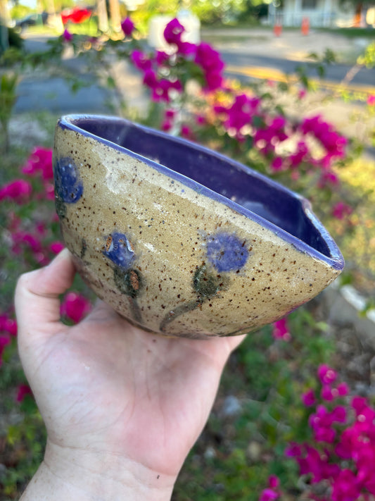 Photo of a speckled ceramic bowl painted with purple thistle flowers on the outside and glazed with a deep purple on the inside.  The bowl is being held in the artist's hand against a blurred background of a green bush with pink flowers.