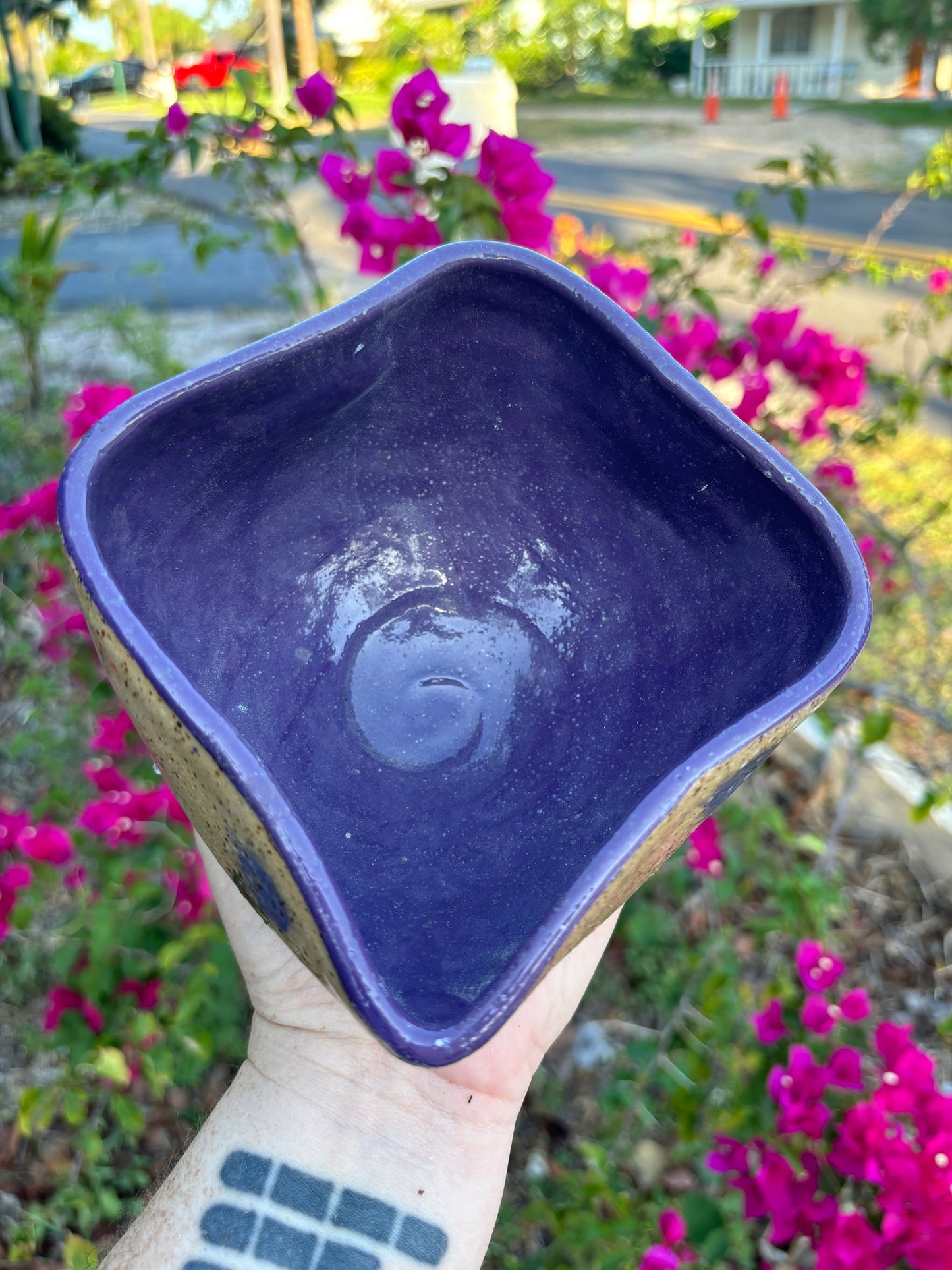 Top down photo of a purple gla\zed bowl in the shape of a yoni.  The bowl is held in the artist's hand against a background of a bush with pink flowers.