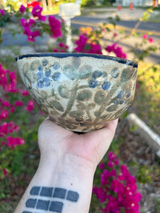 Side view of a ceramic bowl painted with blue cohash berries and leaves.  The bowl is held in the artist's hand against a background of green bushes with pink leaves.