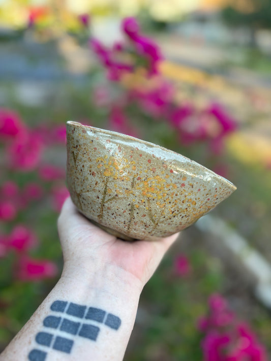 Photo of a brown speckled ceramic bowl with hand painted rue flowers on the sides.  The bowl is held in the artist's hand against a background of bushes with pink flowers.