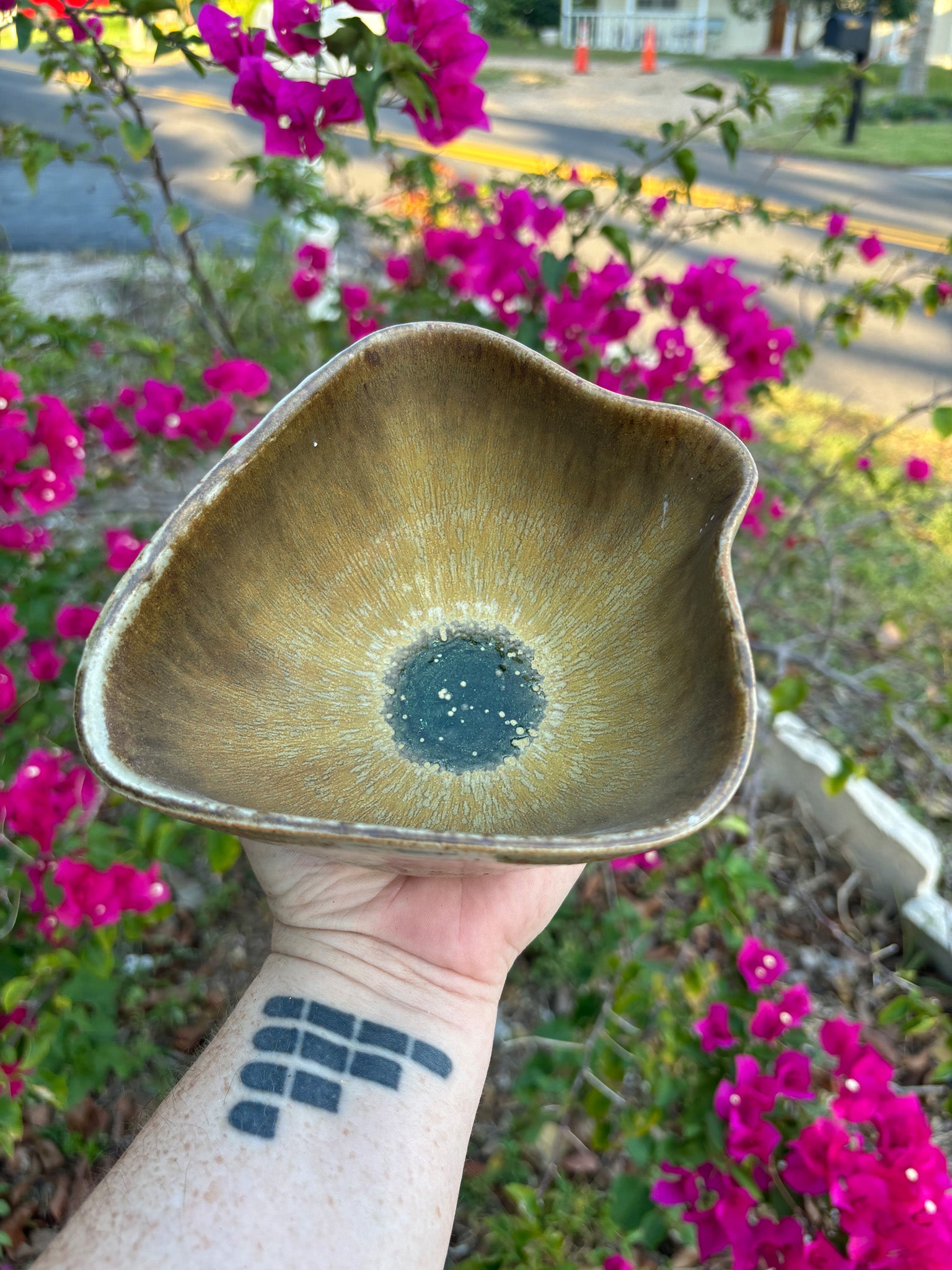 Photo of a yoni-shaped ceramic bowl with buttery glaze that runs elegantly down to the center of the bowl. In the middle, a glassy green pool shines in the sun. The bowl is held in the artist's hand and set against a background of bushes with pink flowers.