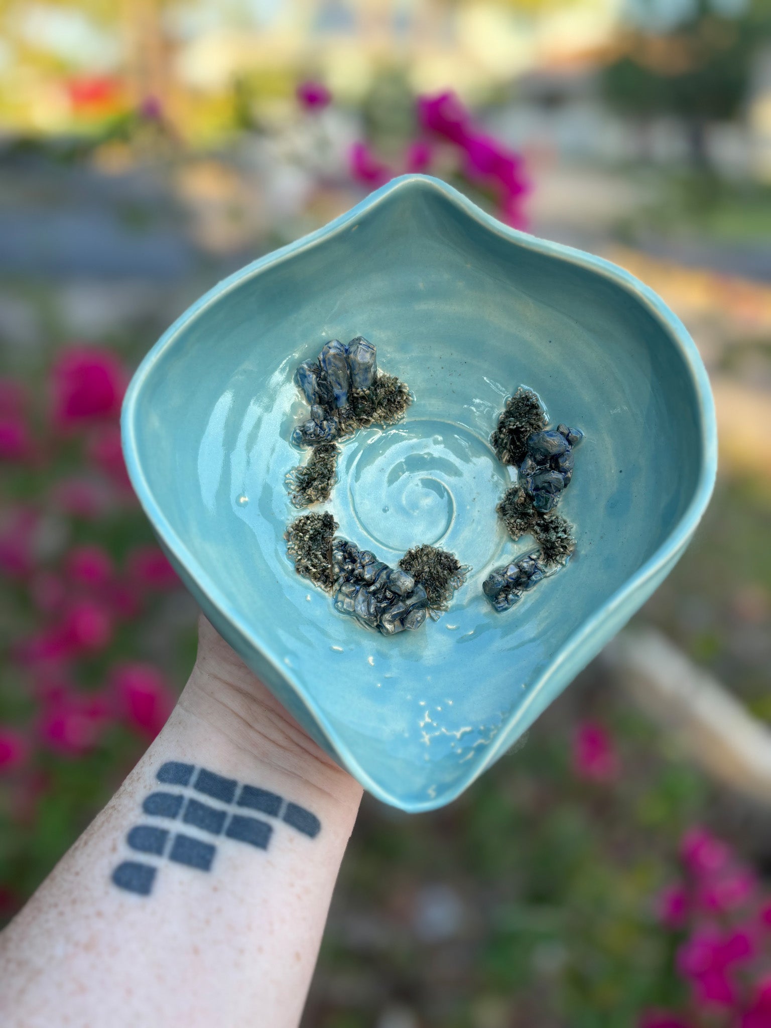 Photo of a yoni shaped vulva bowl in a bright blue green glaze. The center of the bowl has a sacred spiral and there are sculptural crystals growing out of the bottom of the bowl. The clay crystals are painted to look like blue aventurine. The bowl is held in the artist's hand and set against a background of bushes with pink flowers.