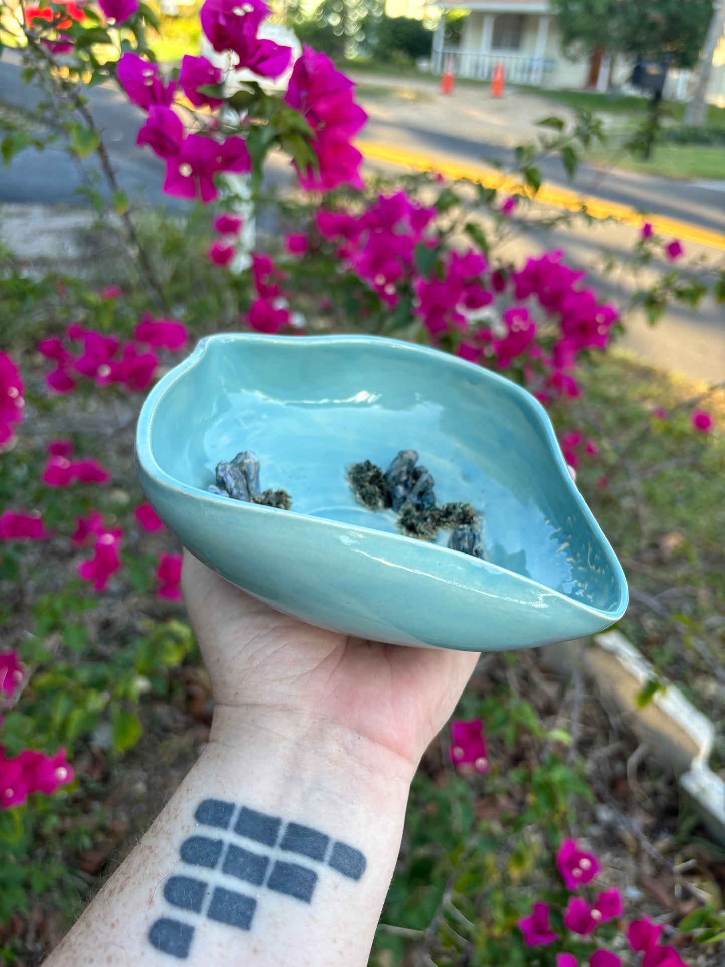 Side view of a yoni shaped vulva bowl in a bright blue green glaze. The center of the bowl has a sacred spiral and there are sculptural crystals growing out of the bottom of the bowl. The clay crystals are painted to look like blue aventurine. The bowl is held in the artist's hand and set against a background of bushes with pink flowers.