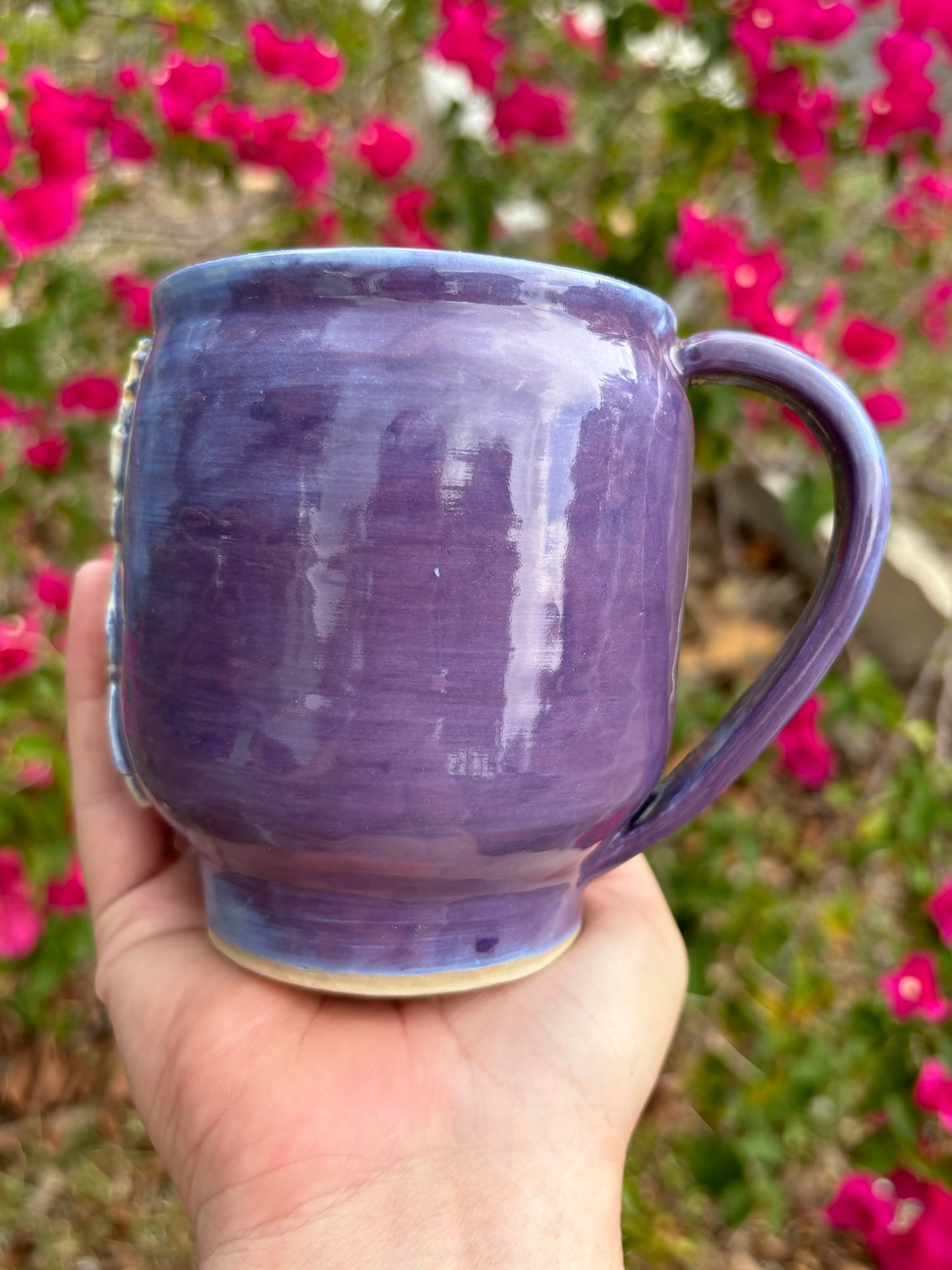 Photo of a large purple mug in the artist's had set against a background of bushes with pink flowers.