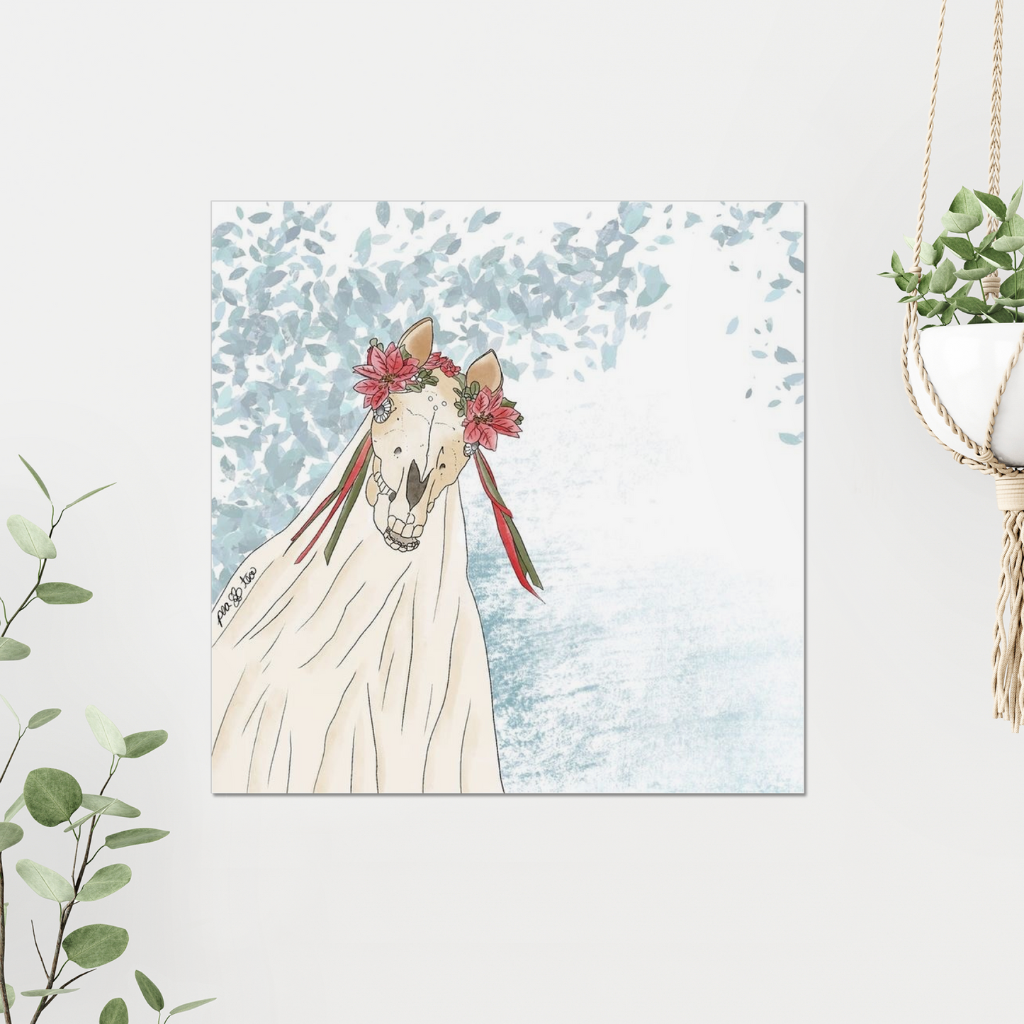 Photo of a watercolor art print of a horse skull on a sheet with colorful winter florals and ribbons on a white wall.  There are green plants framing the print.